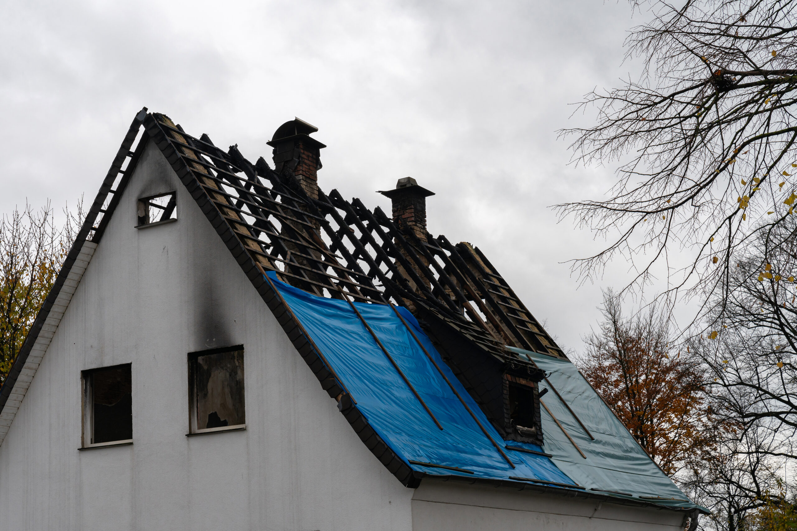 A building shows significant roof damage with a blue tarp covering exposed areas. The cloudy sky adds to the somber atmosphere of the surroundings.